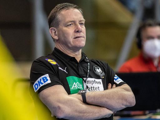 Handball-Bundes-Trainer Alfred Gislason steht in einer Sport-Halle. Er schaut aufmerksam. Das Bild ist gemacht worden beim Olympia-Qualifikations-Spiel Algerien gegen Deutschland.