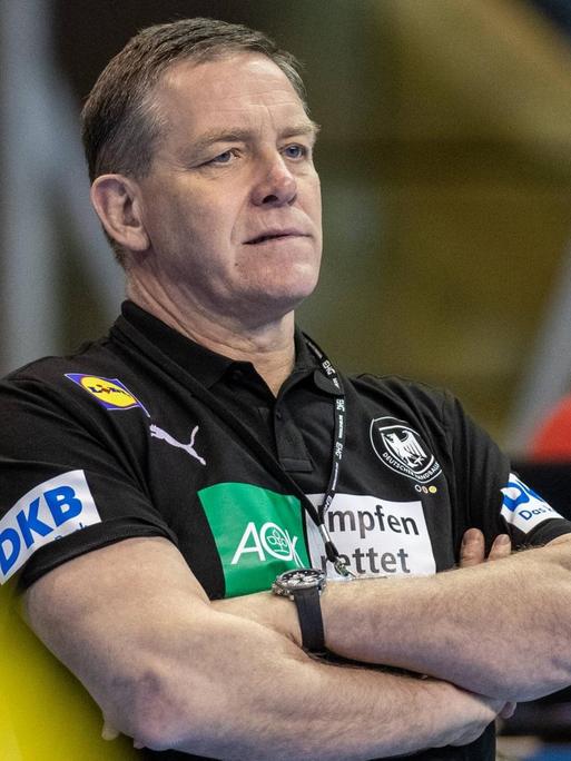 Handball-Bundes-Trainer Alfred Gislason steht in einer Sport-Halle. Er schaut aufmerksam. Das Bild ist gemacht worden beim Olympia-Qualifikations-Spiel Algerien gegen Deutschland.