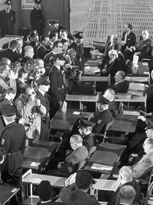 Der erste Auschwitzprozess wird am 20.12.1963 im Plenarsaal der Frankfurter Stadtverordnetenversammlung eröffnet. Das Bild zeigt einen Blick auf die Pressefotografen und Kameraleute, die die Angeklagten aufnehmen. Die Angeklagten sitzen flankiert von Polizisten an den Tischen. Im Hintergrund hängen Pläne des früheren Konzentrationslagers an der Wand.