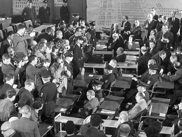 Der erste Auschwitzprozess wird am 20.12.1963 im Plenarsaal der Frankfurter Stadtverordnetenversammlung eröffnet. Das Bild zeigt einen Blick auf die Pressefotografen und Kameraleute, die die Angeklagten aufnehmen. Die Angeklagten sitzen flankiert von Polizisten an den Tischen. Im Hintergrund hängen Pläne des früheren Konzentrationslagers an der Wand.