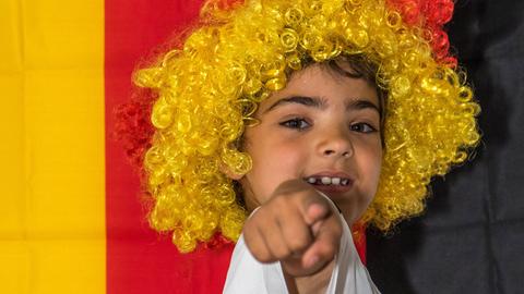 Junger Fan der deutschen Nationalmannschaft in Trikot der Nationalmannschaft mit Perücke in Deutschland-Farben vor deutscher Flagge an Wand