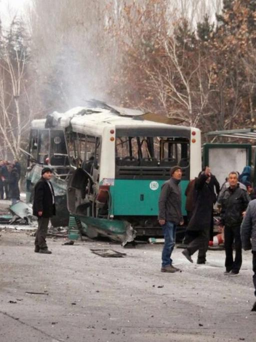 An einer Haltestelle steht ein völlig zerstörter Bus, umringt von Dutzenden Menschen