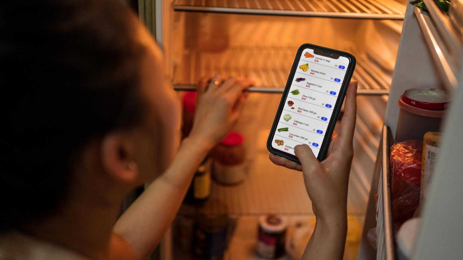 Eine Frau steht vor einem offenen Kühlschrank und bestellt Lebensmittel per App.