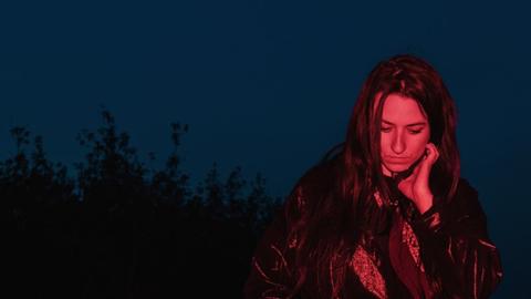 Lucia Cadotsch mit offenen, langen, dunklen Haaren wird rötlich auf einem Feld angestrahlt, während sie vor dunkelblauem Himmel steht - im Hintergrund zeichnet sich ein Wald ab.