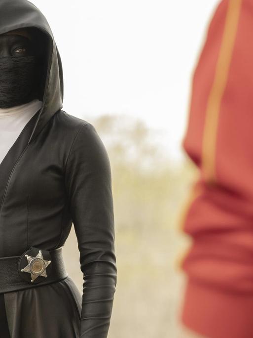 Eine schwarz verhüllte Frau mit Maske trägt einen Sheriffstern an der Hüfte.