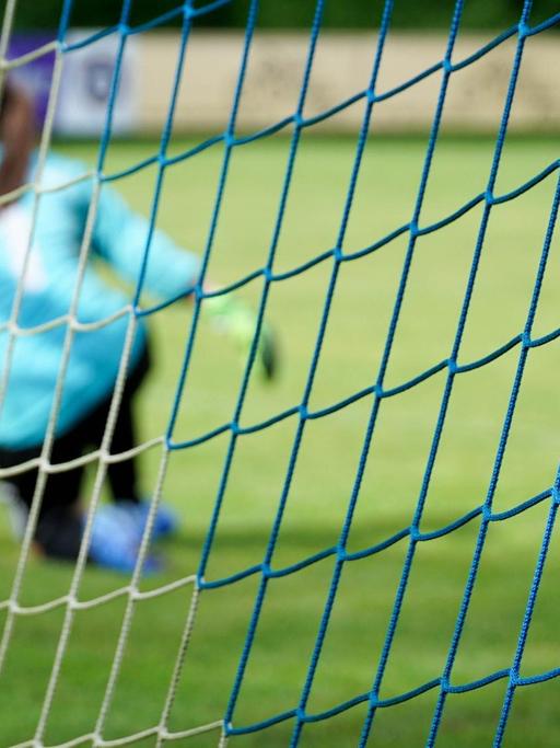 Foto aufgenommen durch das Netz eines Fußballtors: Torfrau in einem hellblauen Trikot kniet auf dem Rasen