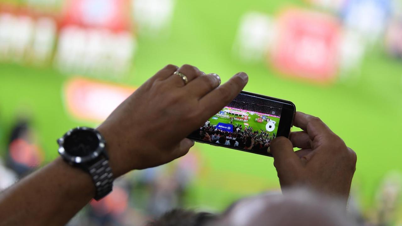 Stadionbesucher sieht das Geschehen auf dem Fußballplatz durch einen Smartphone-Bildschirm