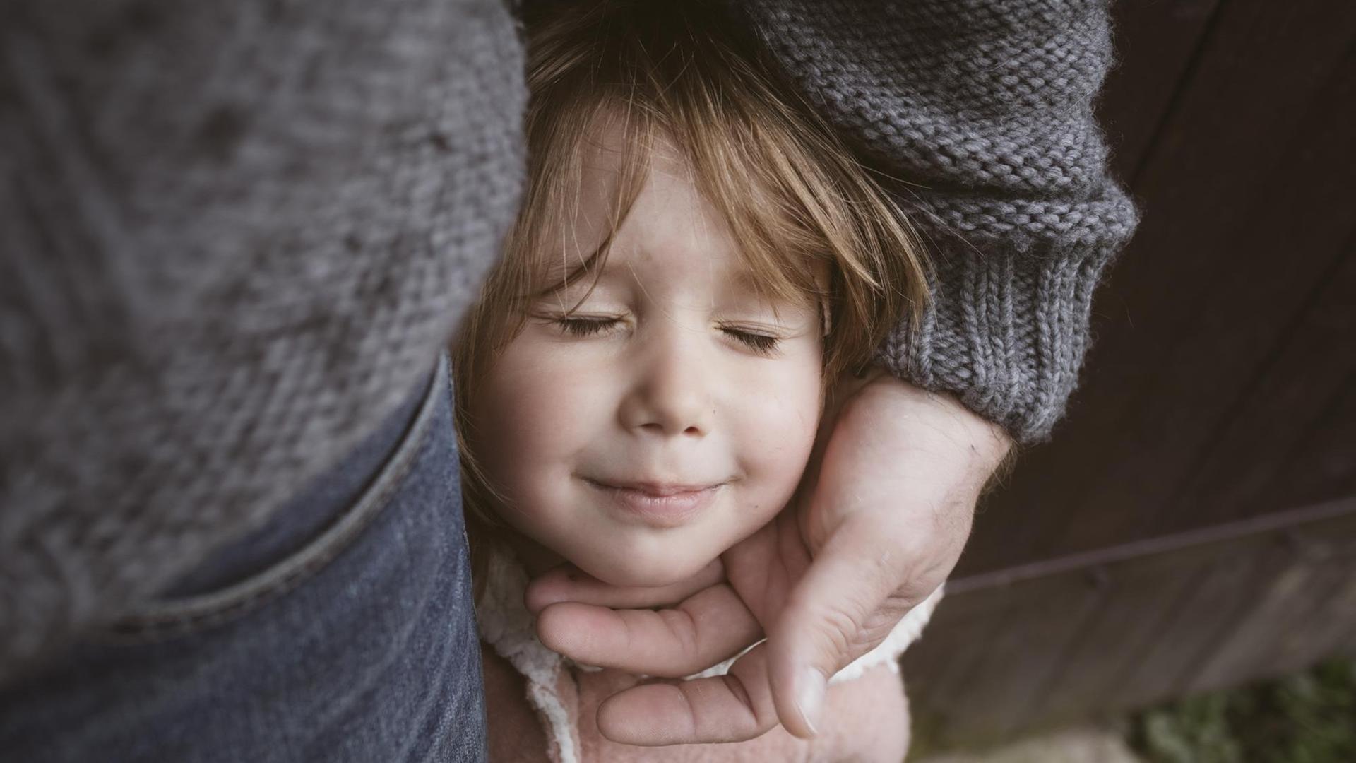 Ein Vater legt beschützend eine Hand um das Gesicht eines Kindes, das Mädchen hält die Augen geschlossen.