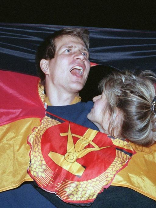 Ein jubelndes Paar hat sich in Berlin, am Tag der Deutschen Einheit in einer DDR-Fahne Platz geschaffen, wo früher das Emblem Hammer und Sichel war – und feiert den historischen Anlass.
