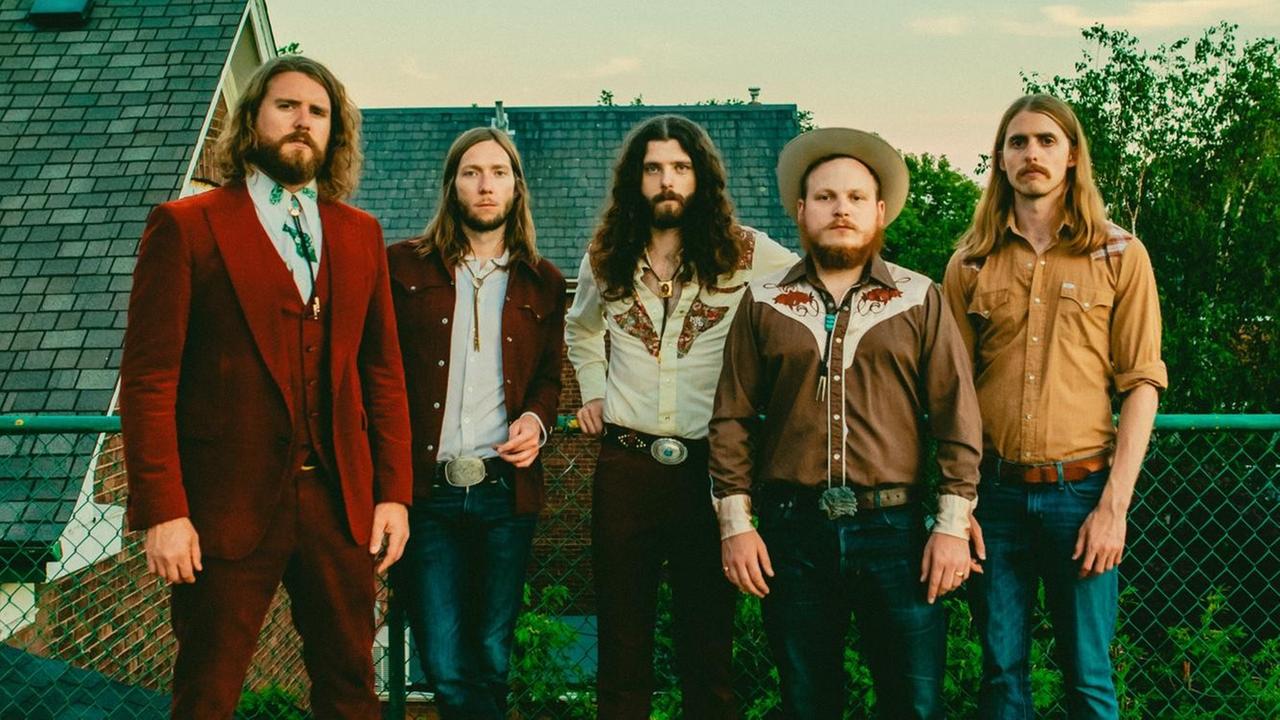 Fünf Männer stehen auf einer Holzterrasse und tragen Vintage-Kleidung, lange Haare und/oder Bart.