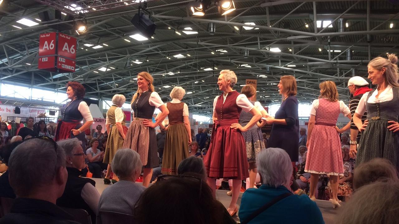 Acht Damen präsentieren klassische Dirndl auf dem Laufsteg der Rentnermesse "Die 66" in München vor vielen älteren Zuschauern.