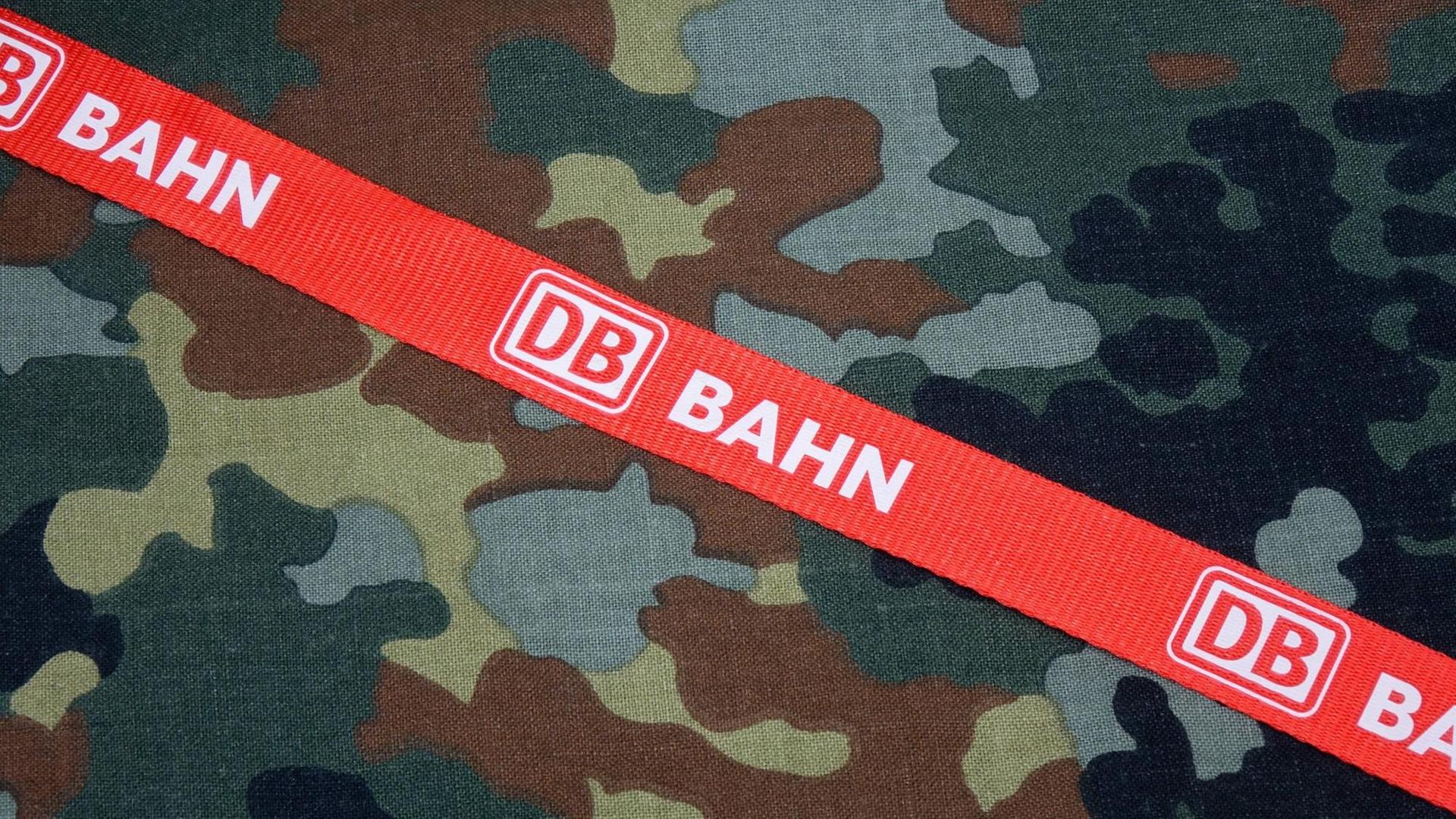 Auf einem Tarnstoff der Bundeswehr, liegt ein Band der Deutschen Bahn mit Logo "DB" und dem Wort "Bahn".