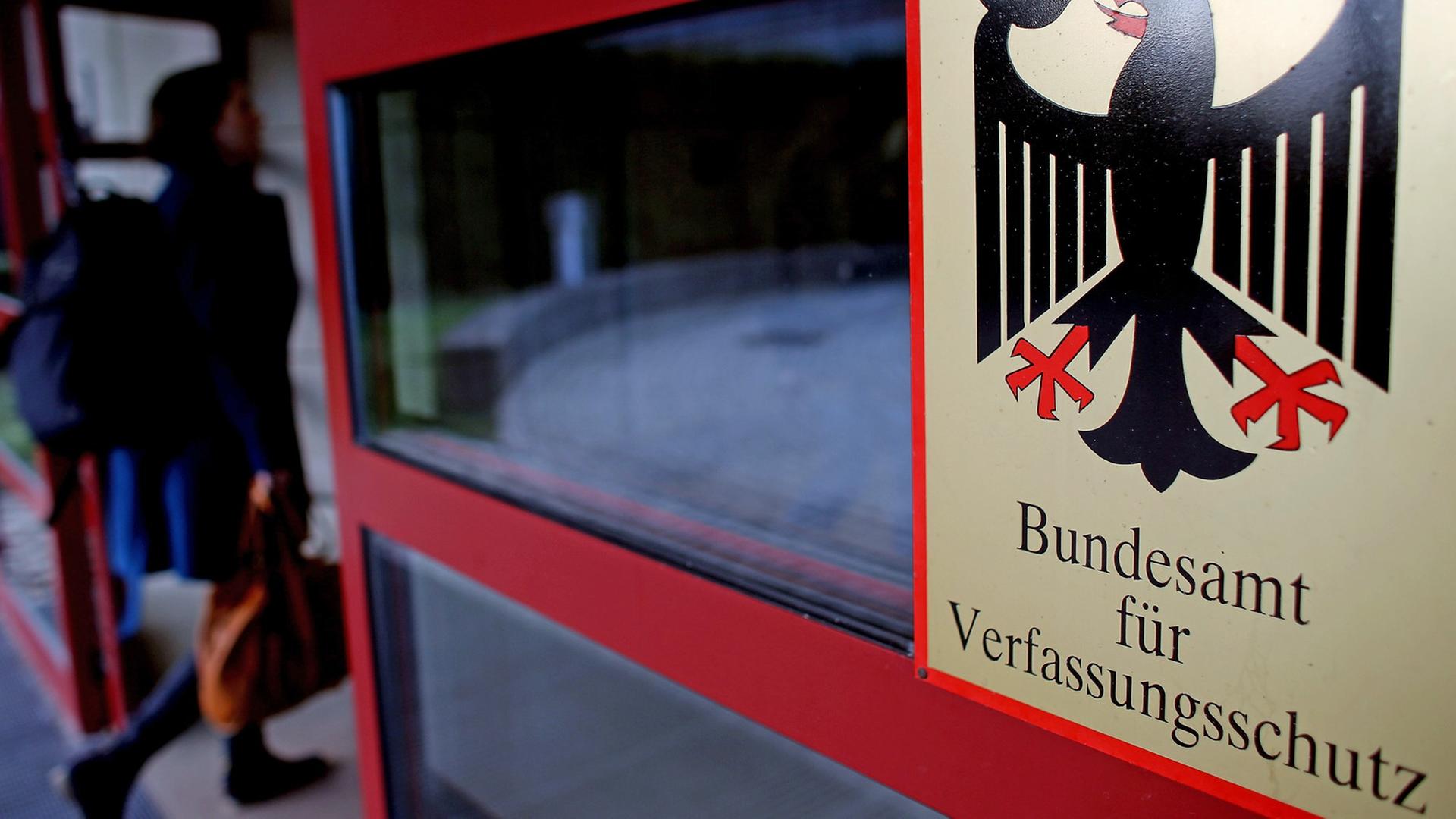 Schild des Bundesamts für Verfassungsschutz mit dem Bundesadler an einer Eingangstür des Gebäudes, durch die eine Frau geht. Schild des Bundesamts für Verfassungsschutz mit dem Bundesadler an einer Eingangstür des Gebäudes, durch die eine Frau geht.