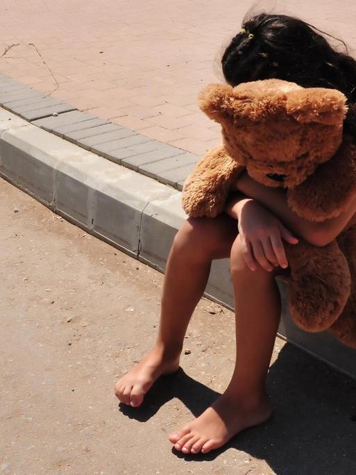 Ein Mädchen sitzt am Straßenrand und vergräbt ihr Gesicht in einem Stofftier, das sie in ihren Armen hält.