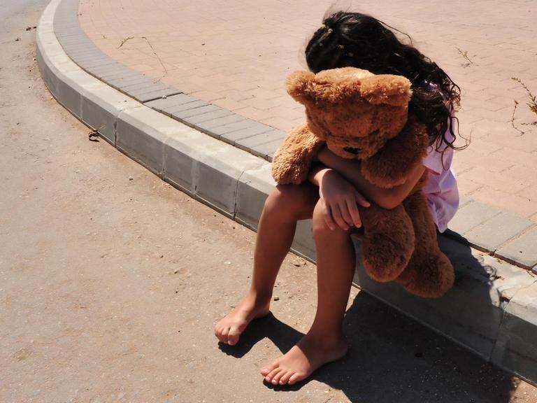 Ein Mädchen sitzt am Straßenrand und vergräbt ihr Gesicht in einem Stofftier, das sie in ihren Armen hält.