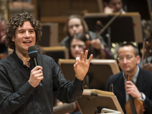 Robin Ticciati steht mit Mikrofon im Vordergrund, im Hintergrund sieht man das Deutsche Symphonie-Orchester Berlin.