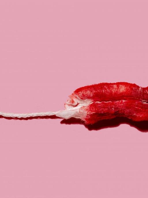 Zu sehen ist ein mit Kunstblut gefärbter Tampon vor einem rosa Hintergrund.