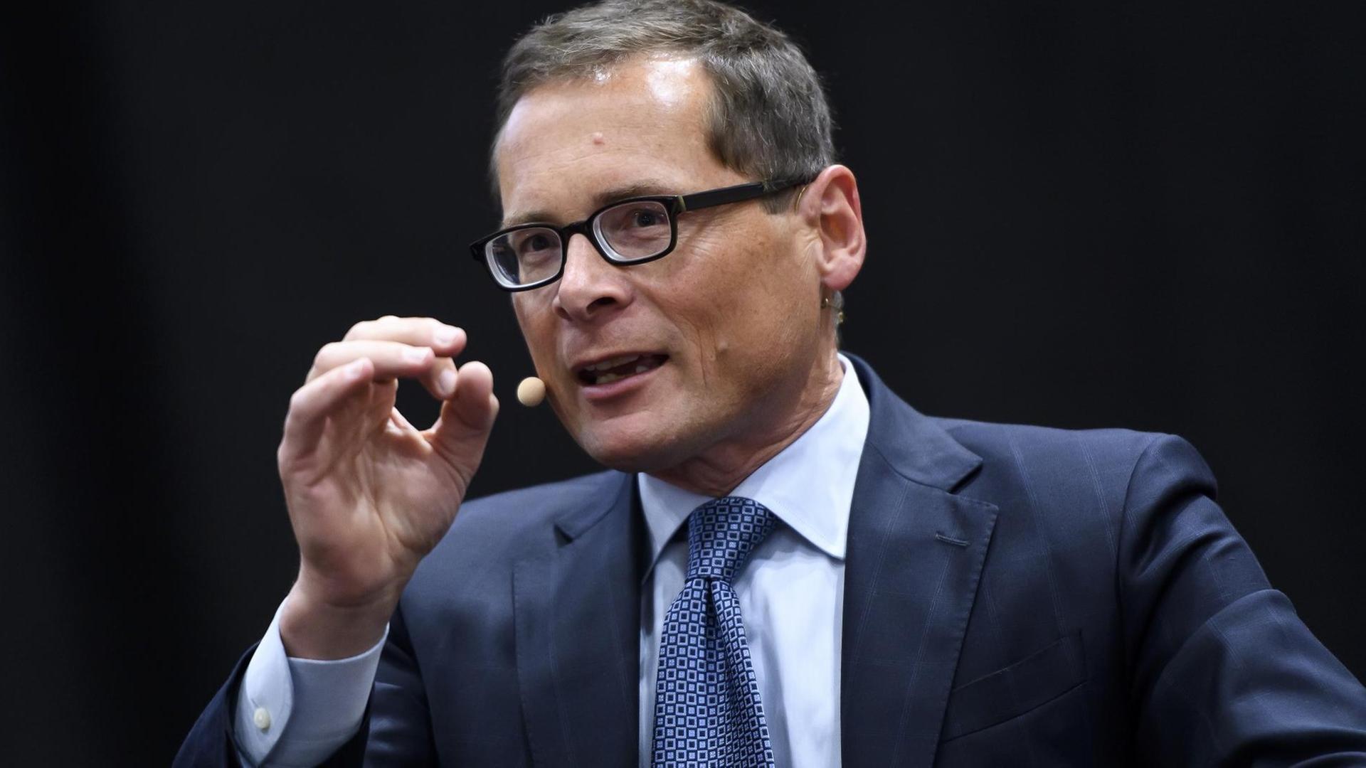 Das Bild zeigt den Chefredakteur Roger Köppel im dunkelblauen Anzug und mit schwarzer, eckiger Brille. Er spricht und gestikuliert dabei mit seiner rechten Hand. Der Hintergrund ist schwarz.