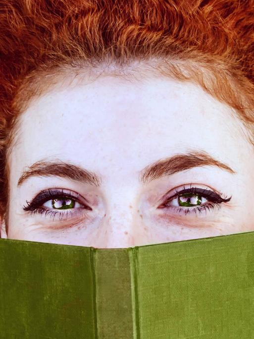 Junge rothaarige Frau bedeckt ihr Gesicht mit einem Buch.