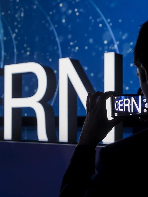 Beleuchteter Schriftzug "Cern" an einer dunklen Wand, rechts im Vordergrund der Schatten einer Person, die den Schriftzug fotografiert