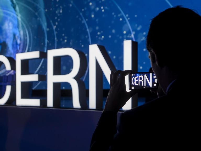 Beleuchteter Schriftzug "Cern" an einer dunklen Wand, rechts im Vordergrund der Schatten einer Person, die den Schriftzug fotografiert