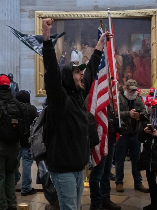 Ein Mob von Trump-Anhängern läuft durch eine Halle im Kapitol. Ein Mann in der Mitte hebt triumphierend die Arme.