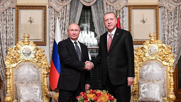 Putin und Erdogan stehen in einem pompös eingerichteten Raum hinter einem Tisch und zwischen zwei mit Gold verzierten Stühlen und schütteln sich die Hände.