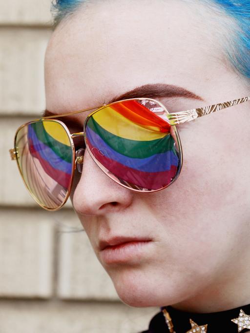 Eine junge Frau mit blau gefärbtem Haar trägt eine Sonnenbrille, in der sich die Regenbogenfahne spiegelt.