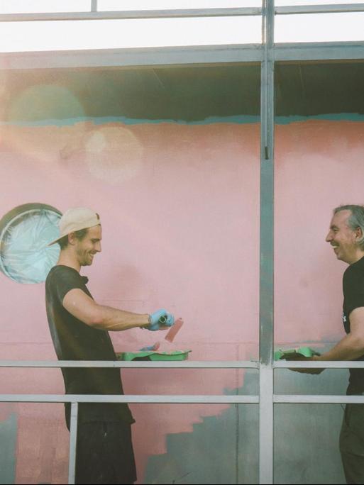 Fynn Kliemann und Olli Schulz stehen an einer zur Hälfte gestrichenen Wand eines Bootes und sind dabei sehr vergnügt.