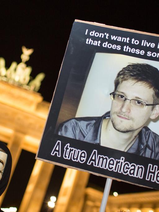Ein Demonstrant der Gruppe Anonymous steht vor dem Brandenburger Tor bei Nacht und hält ein Plakat mit einem Foto von Edward Snowden in die Höhe.