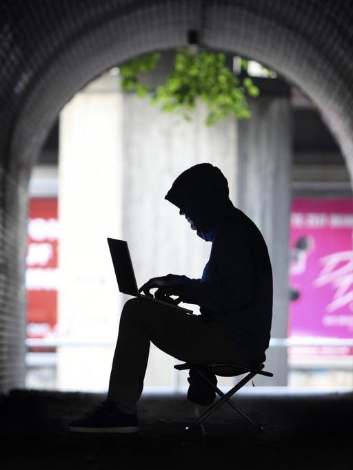 Mann mit Kapuzenpullover sitzt im Schatten eines Tunnels an einem Laptop