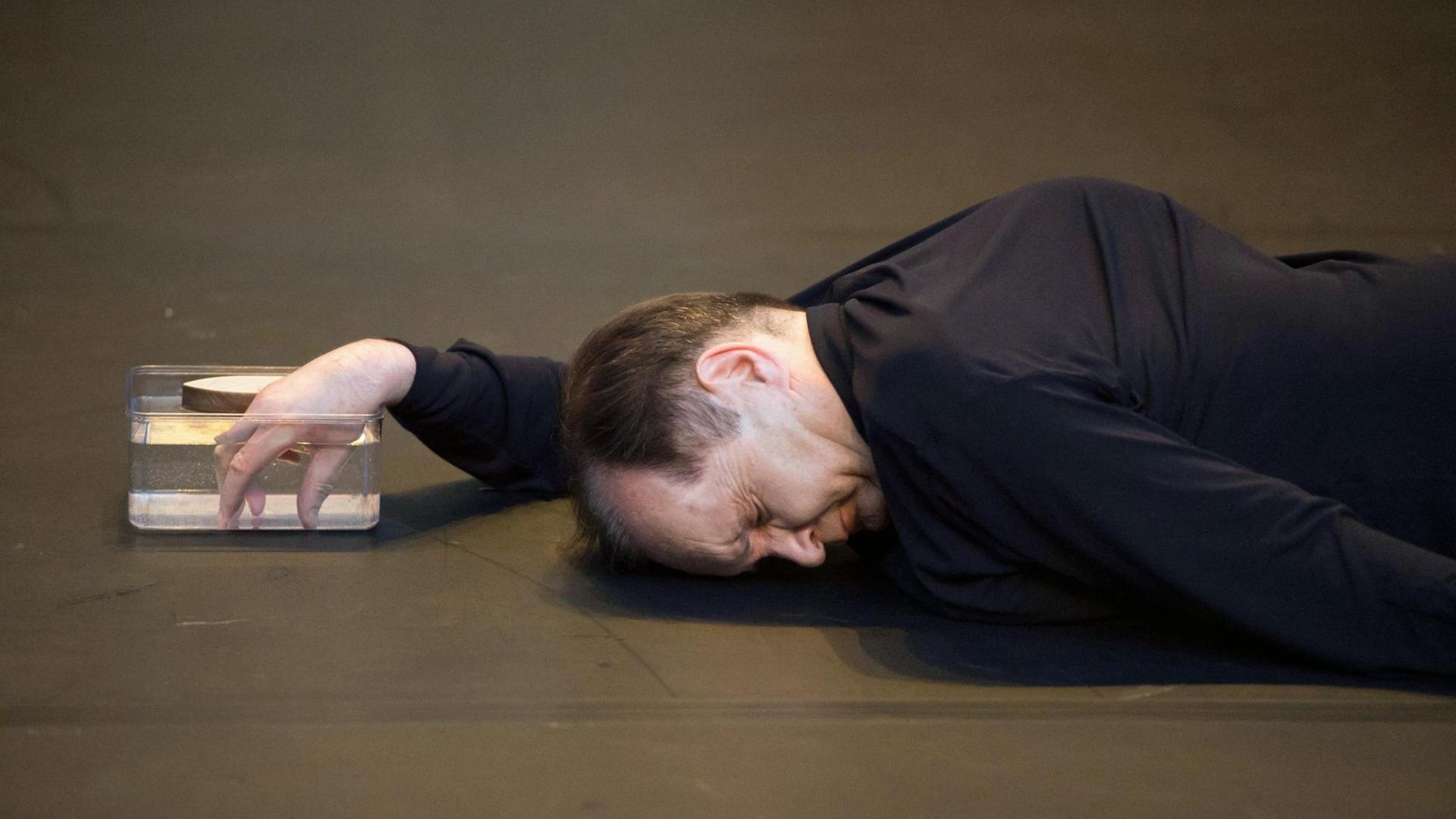 Der Tänzer Raimund Hoghe liegt auf dem Boden und taucht seine rechte Hand in eine durchsichtige Schale, die mit Wasser gefüllt ist.