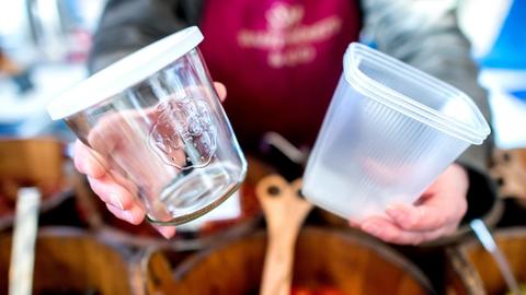 Eine Frau hält auf einem Wochenmarkt ein wiederverwendbares Glas und eine Einwegverpackung aus Plastik in den Händen. Mit dem wachsenden Umweltbewusstsein bei den Verbrauchern setzen die Verkäufer auf Wochenmärkten in Niedersachsen zunehmend auf nachhaltige Verpackungsalternativen.