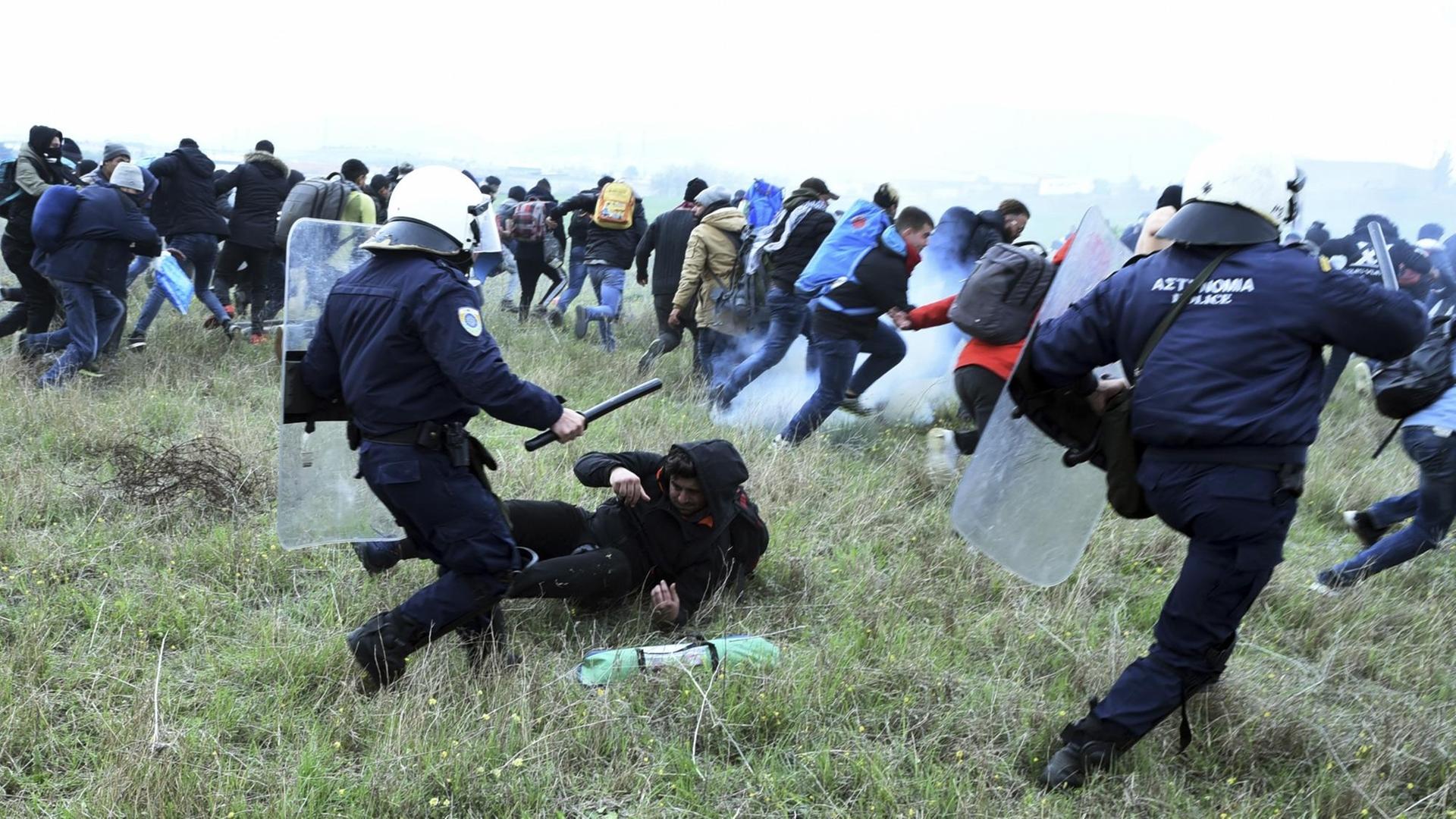 Mit Schlagstöcken bewaffnete Polizisten jagen Flüchtlinge über eine Wiese.