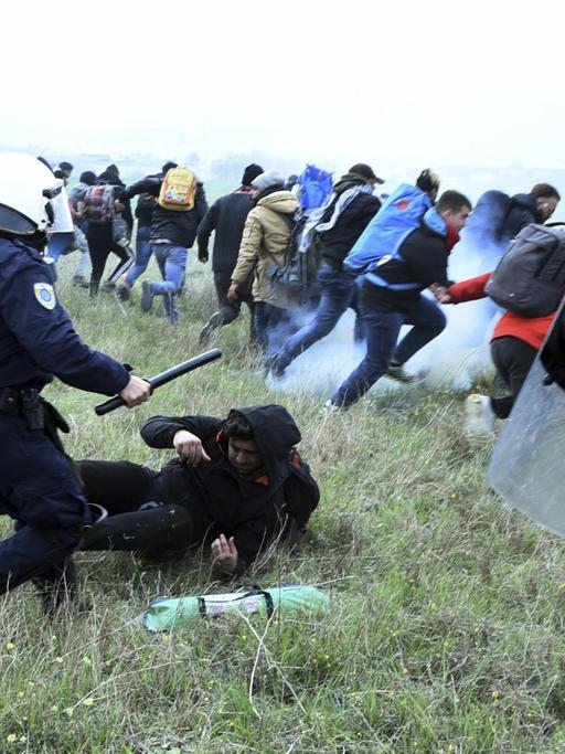 Mit Schlagstöcken bewaffnete Polizisten jagen Flüchtlinge über eine Wiese.