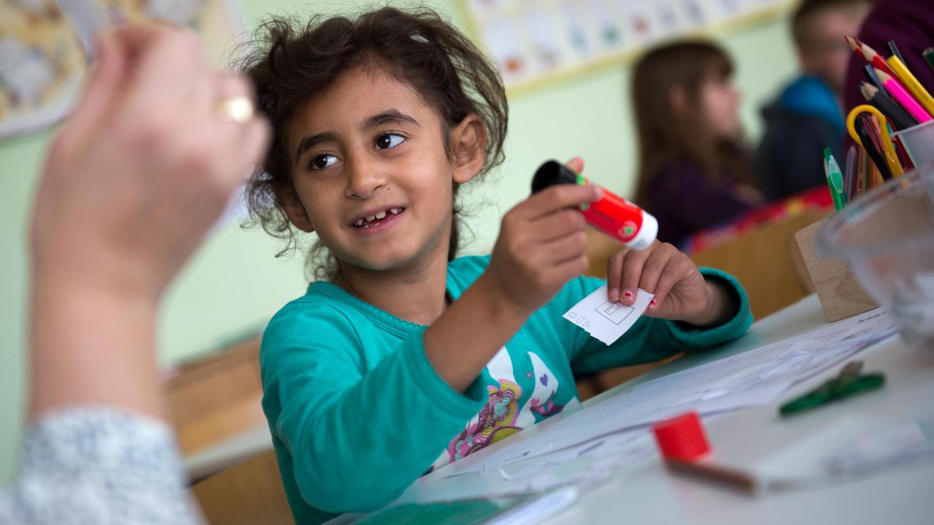 Ein Mädchen mit dunklen Locken arbeitet mit einem Klebestift und lächelt die Lehrerin an.