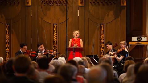 Eine Sängerin in einem roten Kleid steht vor einer braunen Kirchenwand. Links und recht von ihr sitzen Musiker eines Streichquartetts. Vorne im Bild sieht man die Köpfe des Publikums.