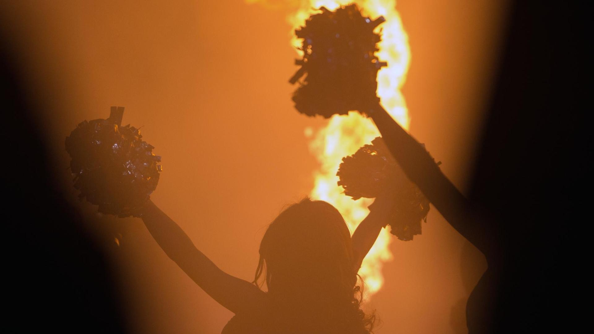Eine Cheerleaderin steht mit ausgestreckten Armen und Pompons an den Händen im Gegenlicht. Im Hintergrund ist eine Feuerfontäne zu sehen.