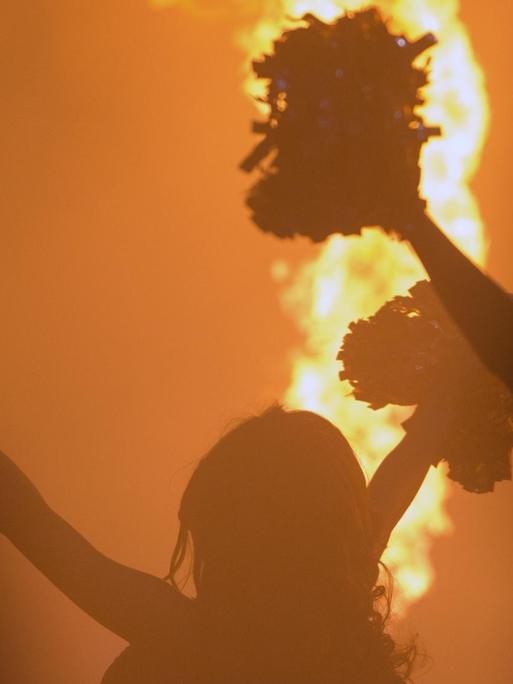 Eine Cheerleaderin steht mit ausgestreckten Armen und Pompons an den Händen im Gegenlicht. Im Hintergrund ist eine Feuerfontäne zu sehen.