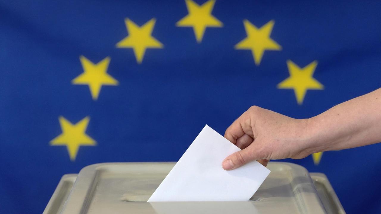 Eine Hand steckt einen Umschlag in eine Wahlurne vor der Europafahne. Symbolfoto.
