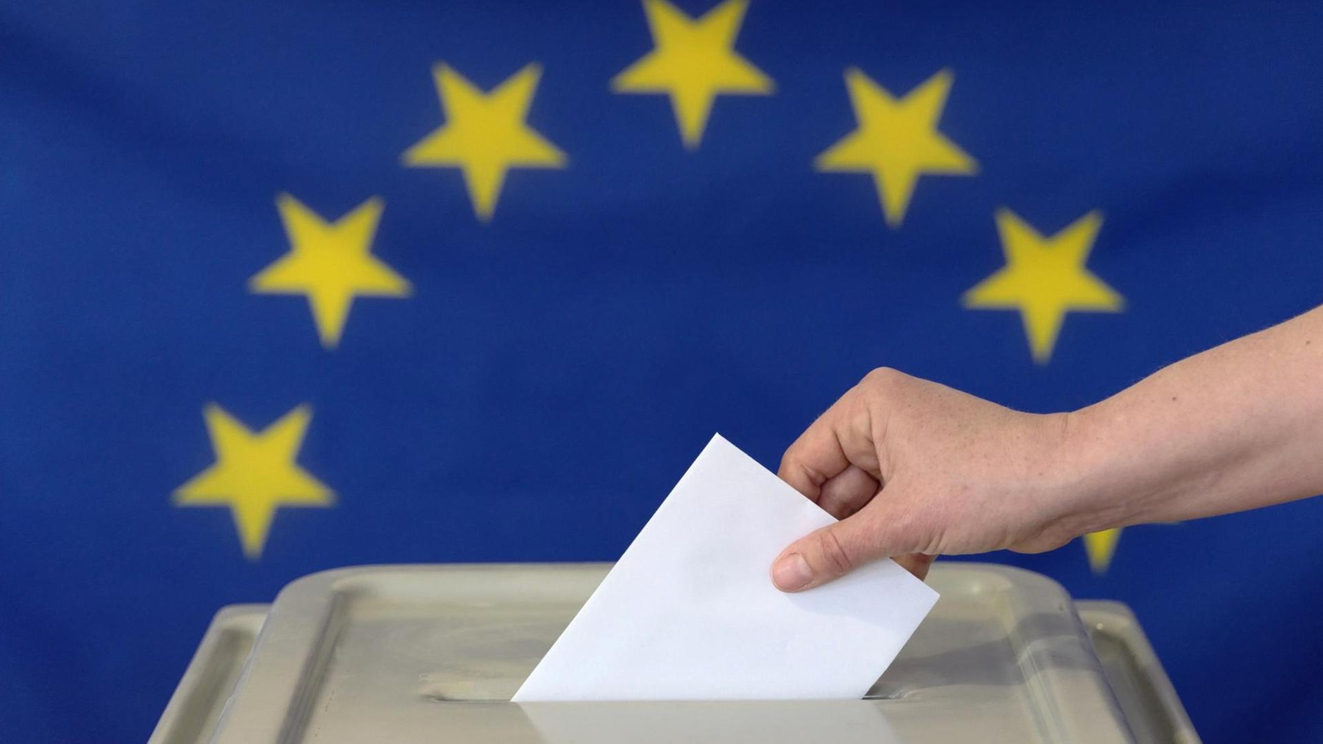 Eine Hand steckt einen Umschlag in eine Wahl-Urne vor der Europa-Fahne.