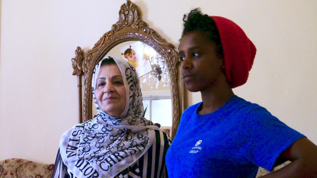 Eine junge Frau (namentlich Zola; im Bild rechts) mit T-Shirt und Mütze steht neben einen älteren Frau, ihrer Dienstherrin (links), in Gewand und Kopftuch