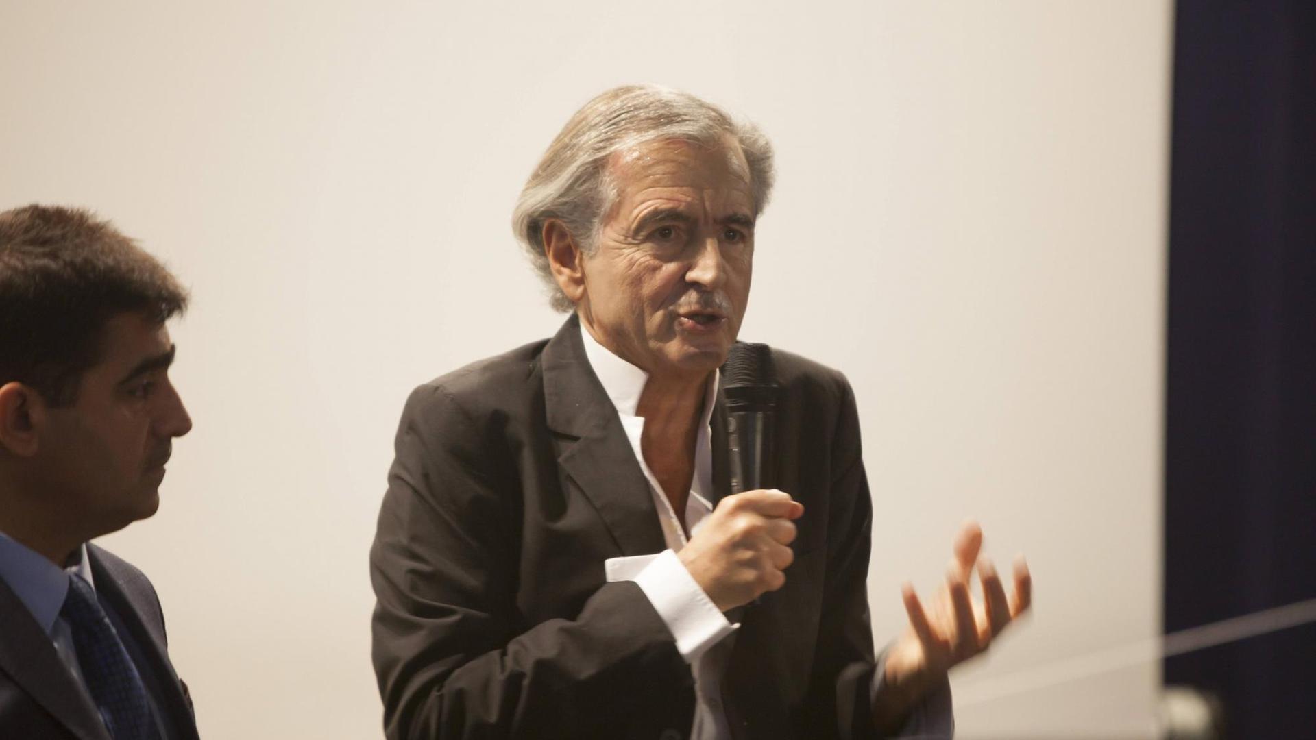 der Philosoph Bernard-Herni Levy steht auf einem Podium und spricht in ein Mikrofon, er gestikuliert mit seiner linken Hand.