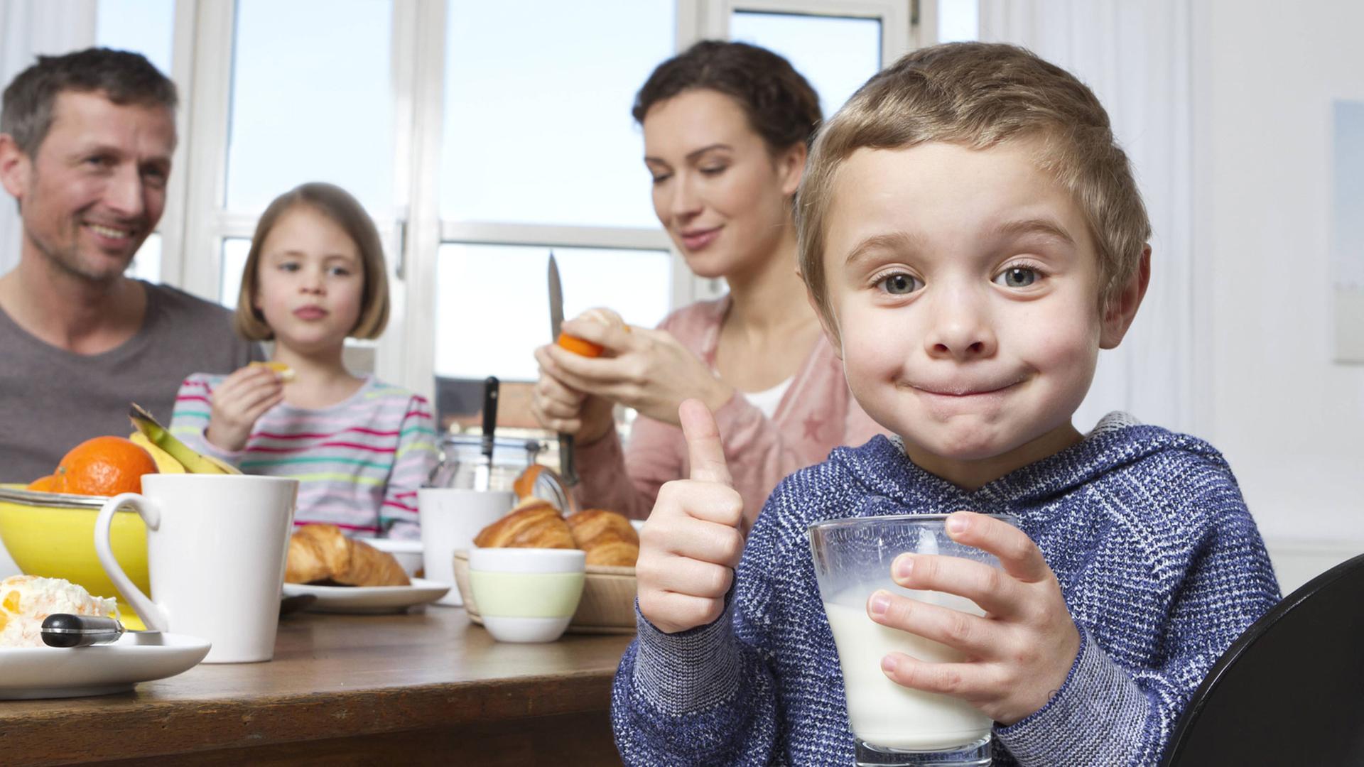 Mutter, Vater, Tochter und Sohn beim Frühstück. Der Sohn hält ein Glas Milch in der Hand und reckt den Daumen in die Kamera.
