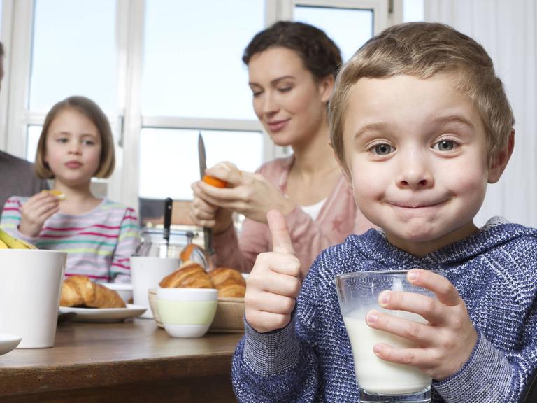 Mutter, Vater, Tochter und Sohn beim Frühstück. Der Sohn hält ein Glas Milch in der Hand und reckt den Daumen in die Kamera.