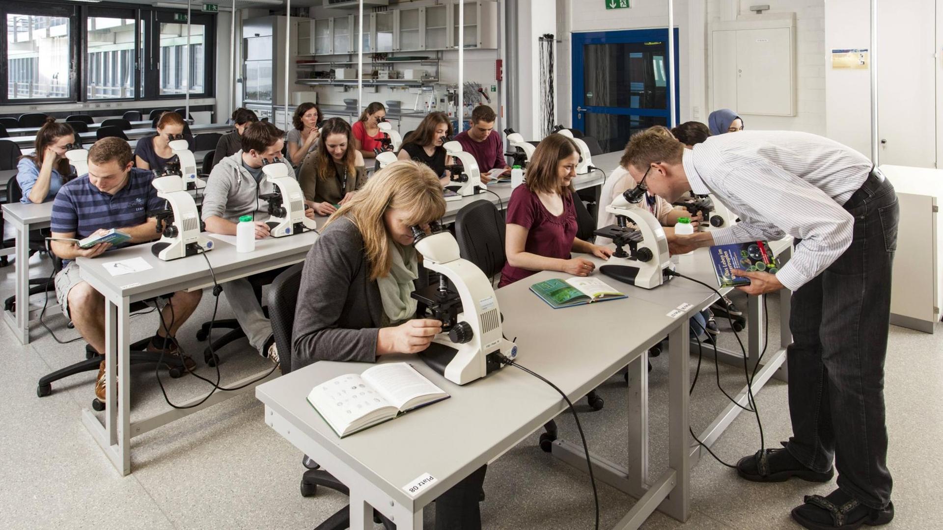 Menschen sitzen in Duisburg in einem Mikroskopierkurs, schauen in die Mikroskope, ein Lehrer hilft.