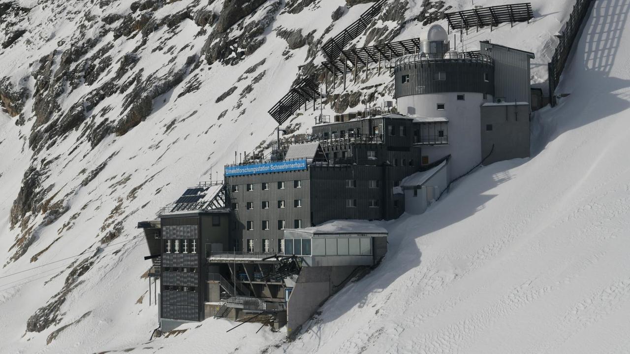 Ein Farbfoto zeigt einen terrassierten, grau verkleideten Gebäudekomplex inmitten eines steil abfallenden, schneebedeckten Felsmassivs 