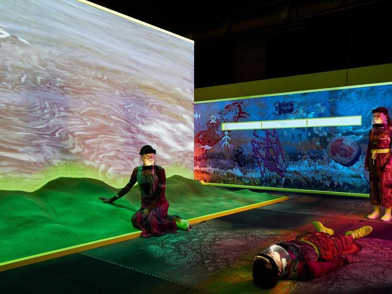 Großformatige farbige Plakate zeigen eine futuristische Landschaft in der drei Menschen mit Schutzkleidung. Auf der Bühne die Schauspieler (v.l.n.r.) Marie Groothof, Thomas Hauser und Ixchel Mendoza Hernandez