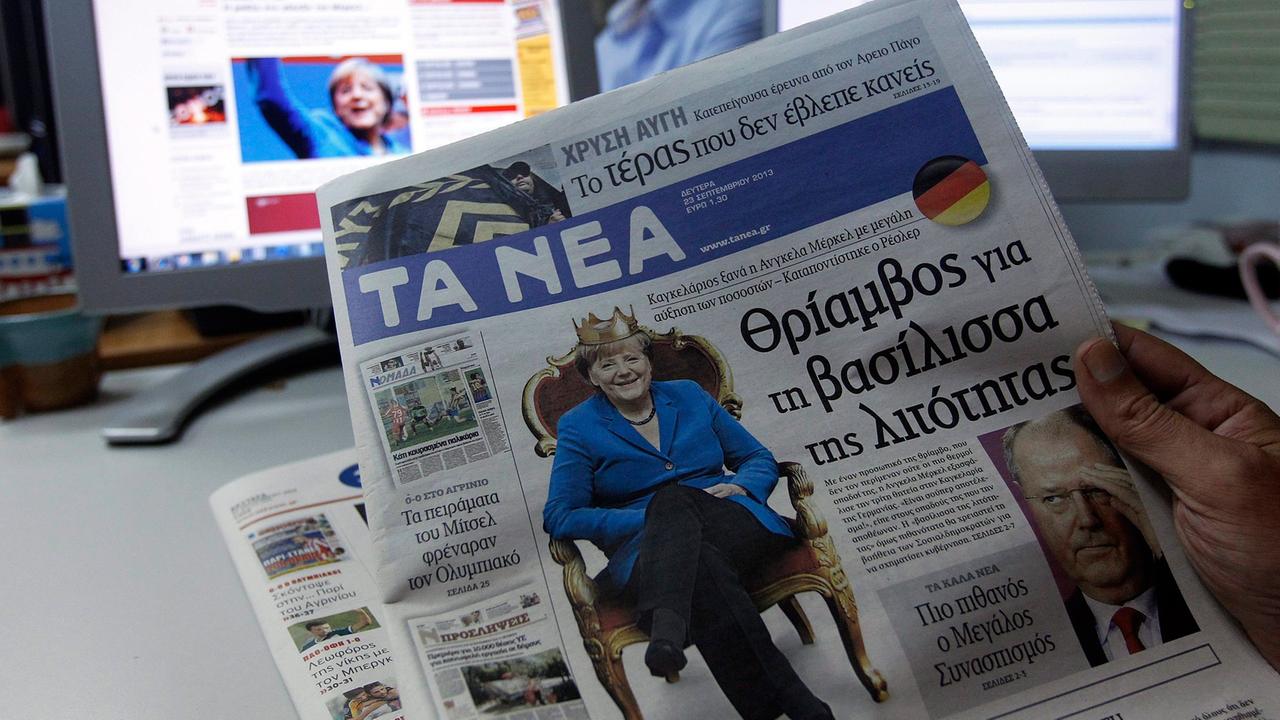 Ein griechischer Journalist hält eine Tageszeitung in der Hand, auf der Bundeskanzlerin Merkel auf einem Thron sitzt.
