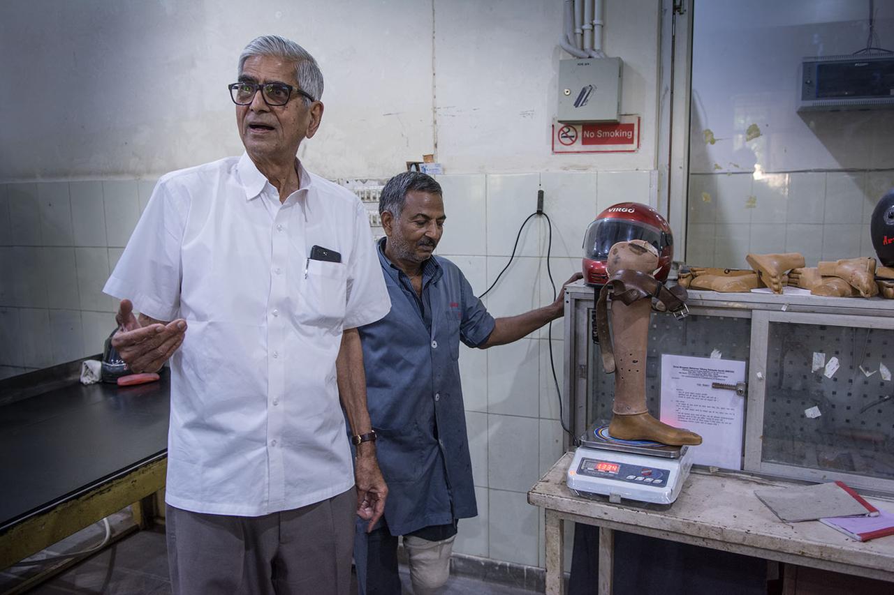 Dr. Mehta steht mit einem Mitarbeiter in der Werkstatt, beide schauen in die Kamera, im Hintergrund eine Prothese auf einer Waage